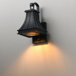 A black metal outdoor wall sconce with a warm yellow light glowing from within, mounted on a plain light gray wall.