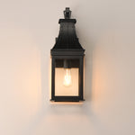 A black metal lantern-style wall sconce with a warm glowing Edison bulb, mounted on a plain off-white wall.