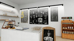 A modern coffee shop interior with a white counter, wooden shelving, display racks of packaged goods, and three large black chalkboard-style wall menus featuring coffee-themed illustrations and text.