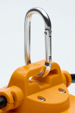 Close-up of a silver metal carabiner clipped onto an orange plastic mounting bracket, with part of a black cable visible against a light gray background.