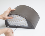 A person's hands are shown installing or adjusting a brown outdoor LED light fixture with a clear lens, using a screwdriver against a clean white background.