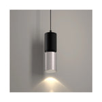 A modern cylindrical pendant light with a matte black upper section and a brushed nickel lower shade, shown illuminated against a plain gray wall.