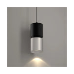 A modern cylindrical pendant light with a matte black upper section and a brushed nickel lower shade, suspended against a neutral gray wall with a warm light beam casting downward.