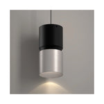 A modern cylindrical pendant light with a matte black top section and a brushed nickel metal lower shade, suspended against a neutral gray wall with a warm light glowing downward.