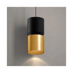 A modern cylindrical pendant light with a matte black upper shade and a brushed gold lower shade, suspended against a neutral gray wall with a warm light glowing downward.