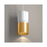 A modern cylindrical pendant light with a matte white upper section and a brushed gold lower section, shown hanging against a neutral gray wall with a warm light glow below.