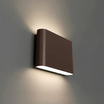 A modern rectangular wall sconce light fixture in a bronze finish, mounted on a gray wall and casting a warm light downward.