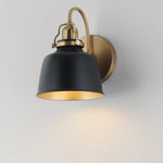 A modern wall-mounted sconce with a matte black conical shade, a warm brass/gold-toned curved arm and wall plate, shown lit against a plain light gray wall.