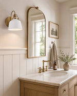 A styled bathroom interior featuring a light wood vanity with a white marble countertop and an antique brass faucet, beneath a large arched gold-framed mirror. A white wall sconce, white wainscoting, a beige towel, and a small potted plant are also visible.