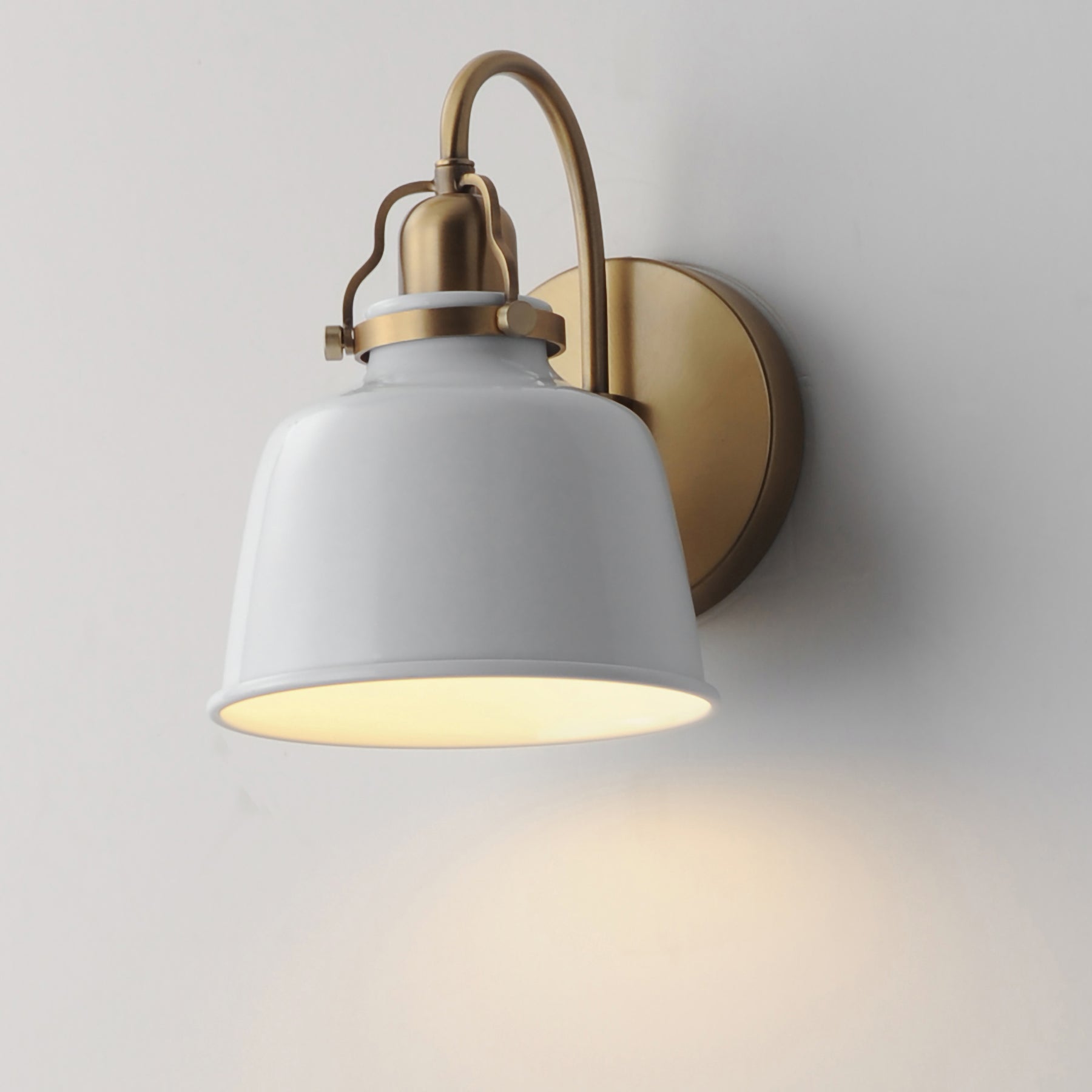 A modern wall sconce light fixture with a white metal bell-shaped shade and a brushed brass/gold curved arm and circular wall mount, shown lit against a plain light gray wall.