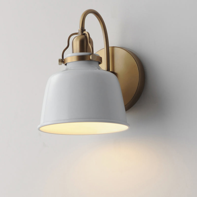 A modern wall sconce light fixture with a white metal bell-shaped shade and a brushed brass/gold curved arm and circular wall mount, shown lit against a plain light gray wall.