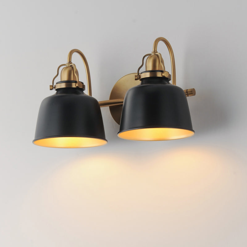 A two-light wall sconce with matte black metal shades and warm brass/gold-toned hardware, shown lit against a plain light gray wall.