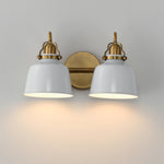 A two-light wall sconce with white bell-shaped shades and antique brass hardware, shown mounted on a light gray wall with warm light glowing from the bulbs.