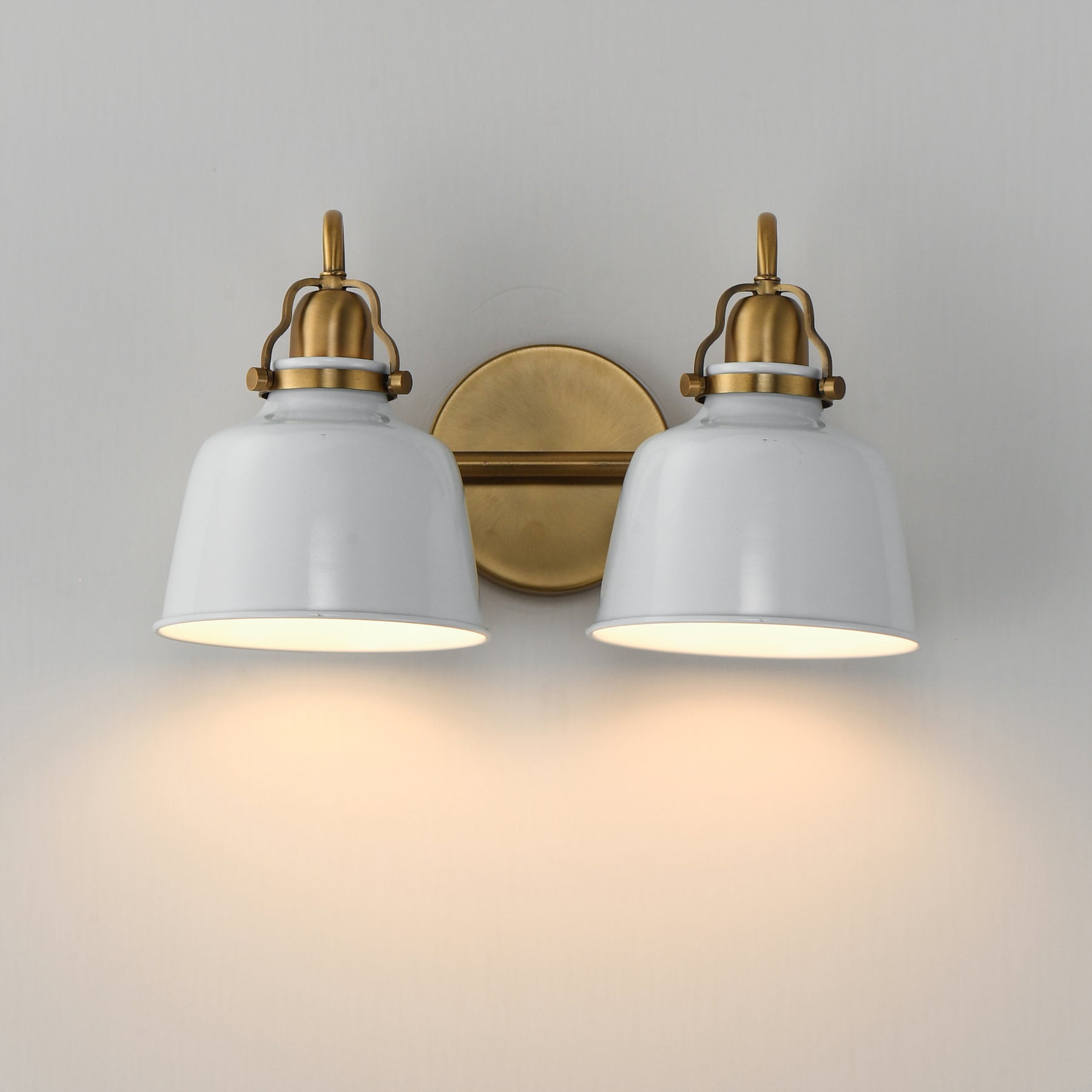 A two-light wall sconce with white bell-shaped shades and antique brass hardware, shown mounted on a light gray wall with warm light glowing from the bulbs.