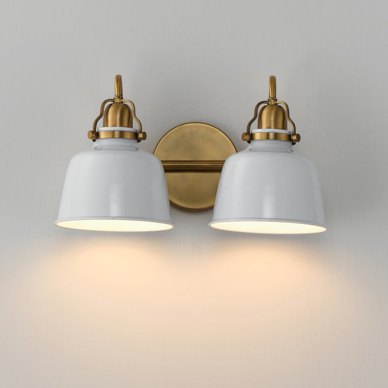 A two-light wall sconce with white bell-shaped shades and antique brass hardware, shown mounted on a light gray wall with warm light glowing from the bulbs.