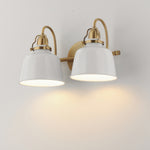 A two-light wall sconce with white bell-shaped shades and a brushed gold/brass metal frame, mounted on a plain light wall and casting a warm glow downward.