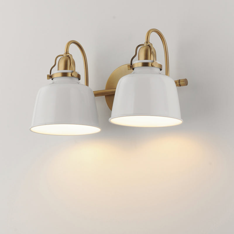 A two-light wall sconce with white bell-shaped shades and a brushed gold/brass metal frame, mounted on a plain light wall and casting a warm glow downward.