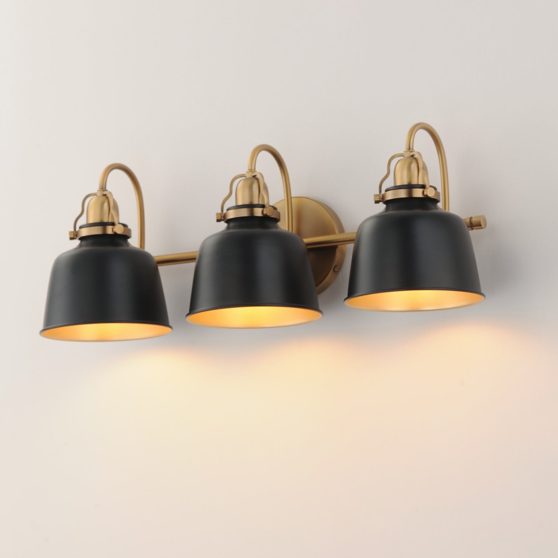 Three-light wall sconce with matte black bell-shaped shades and warm brass hardware, shown mounted on a plain light wall with the lights turned on and casting a warm glow.