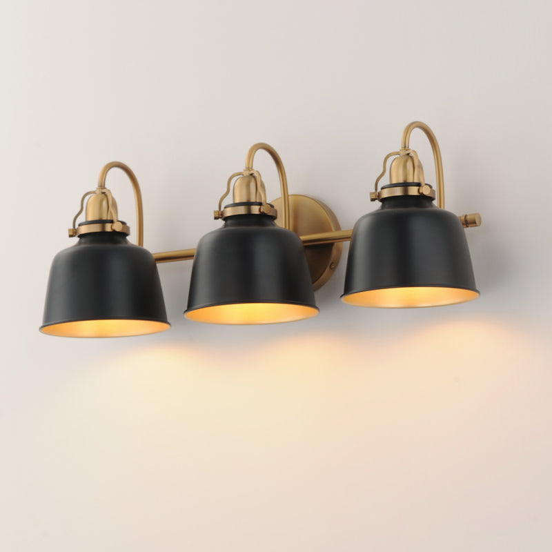 Three-light wall sconce with matte black bell-shaped shades and warm brass hardware, shown mounted on a plain light wall with the lights turned on and casting a warm glow.