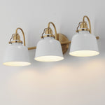 Three-light wall sconce with white bell-shaped glass shades and brushed brass hardware, shown mounted on a light wall with warm illumination.