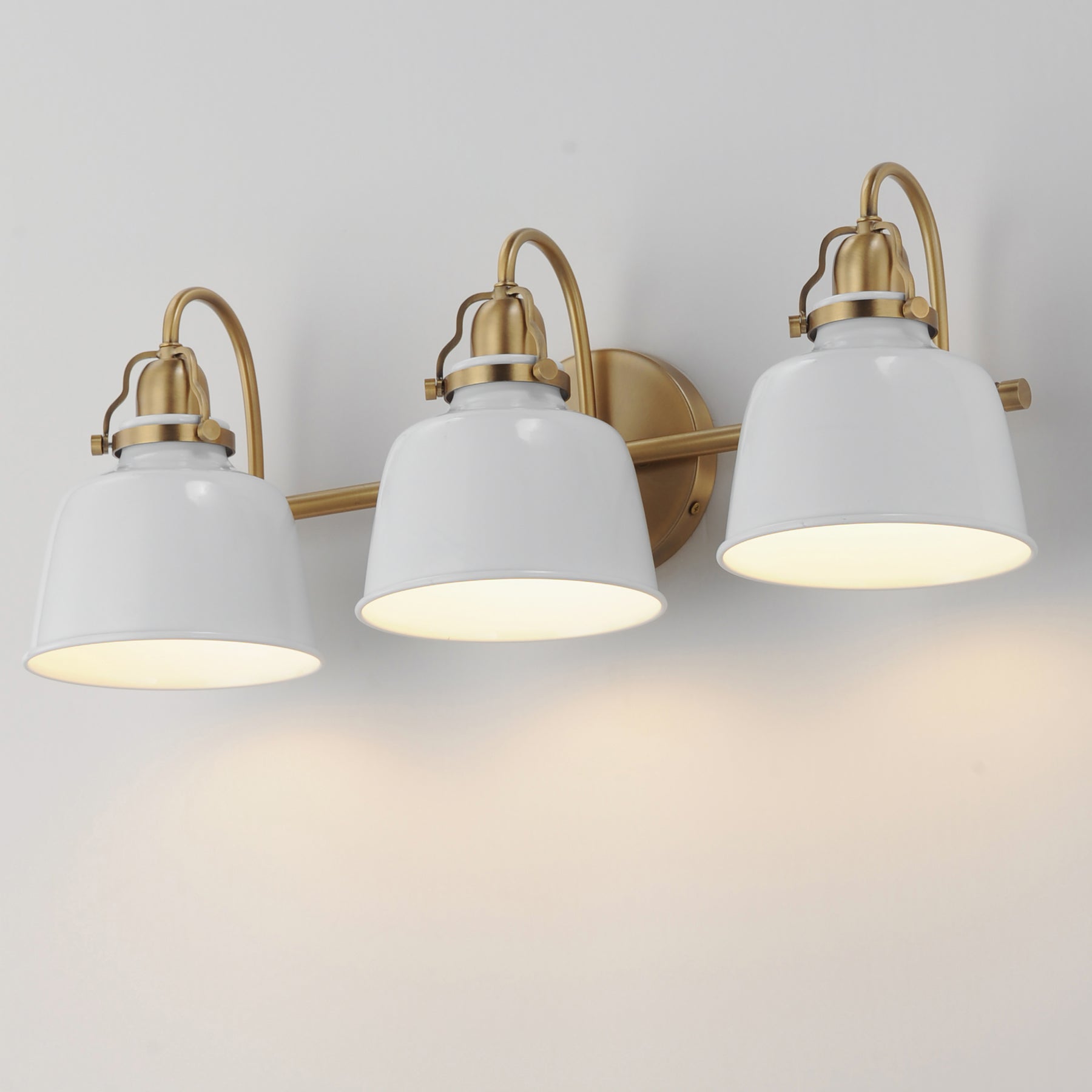 Three-light wall sconce with white bell-shaped glass shades and brushed brass hardware, shown mounted on a light wall with warm illumination.