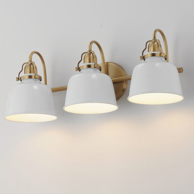 Three-light wall sconce with white bell-shaped glass shades and brushed brass hardware, shown mounted on a light wall with warm illumination.