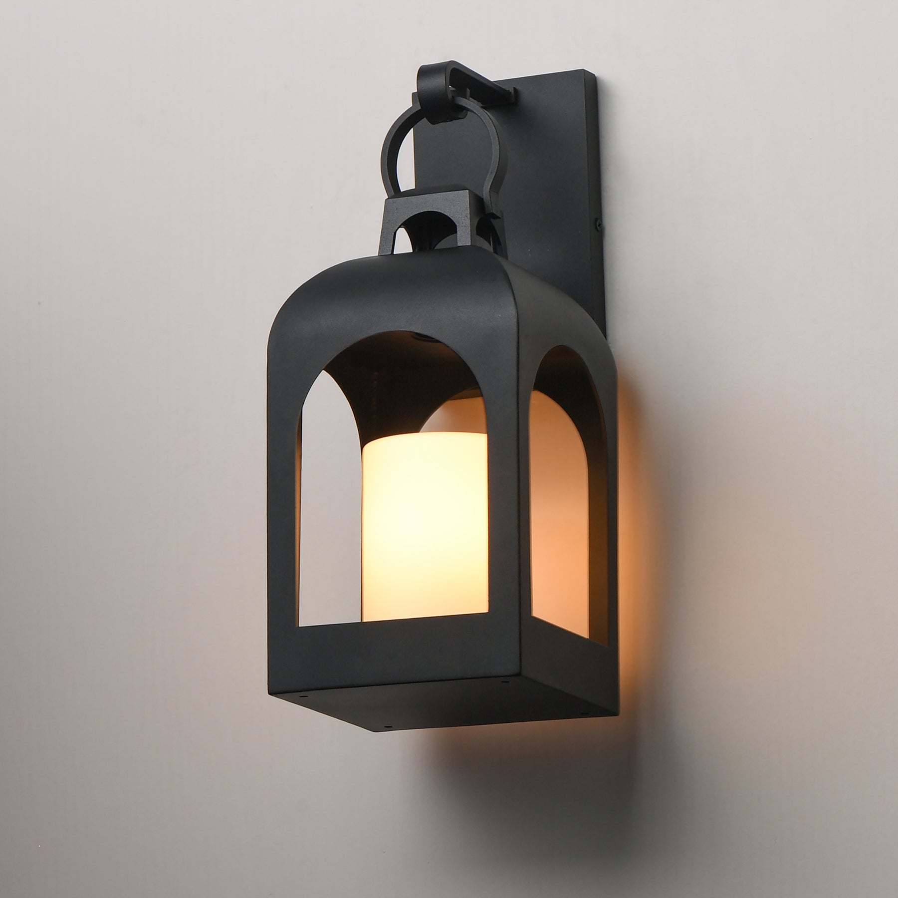 A black metal wall sconce lantern light with a warm glowing candle-style bulb inside, mounted on a plain light gray wall.