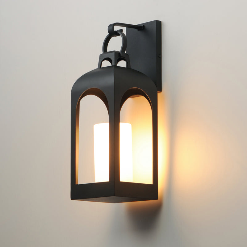 A black metal lantern-style wall sconce with a warm glowing candlelight bulb inside, mounted on a plain light wall.