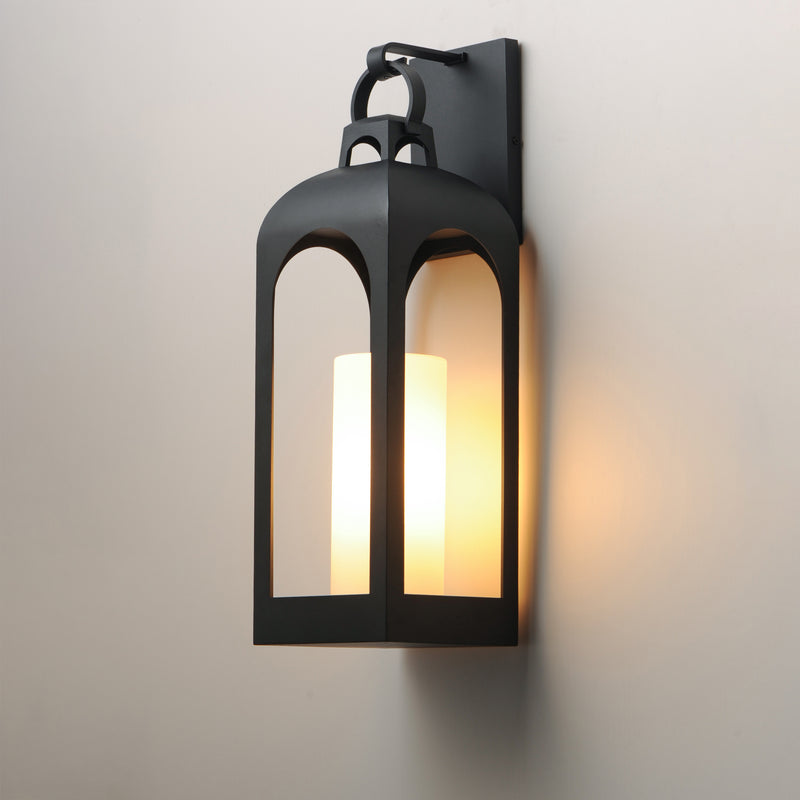 A black metal lantern-style wall sconce with a warm white candle-style light inside, mounted on a plain light gray wall.