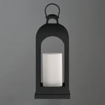A black metal lantern-style candle holder with a white cylindrical candle inside, centered against a solid gray background.