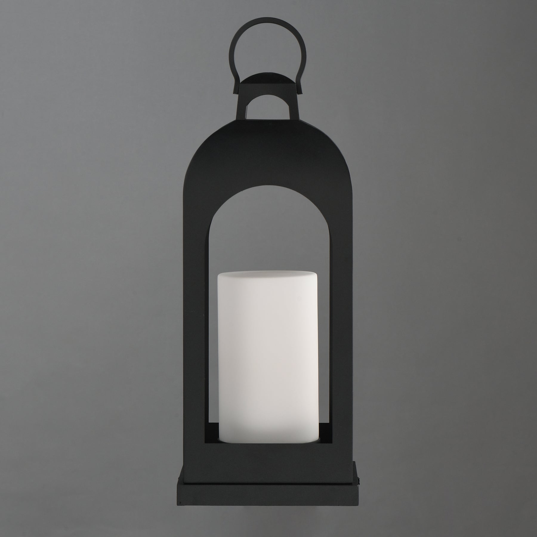 A black metal lantern-style candle holder with a white cylindrical candle inside, centered against a solid gray background.