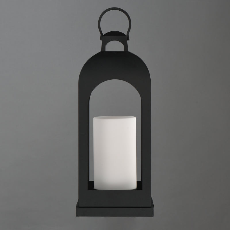 A black metal lantern-style candle holder with a white cylindrical candle inside, centered against a solid gray background.