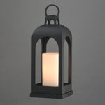 A black metal lantern-style outdoor light with a glowing warm white cylindrical candle inside, shown against a plain gray background.