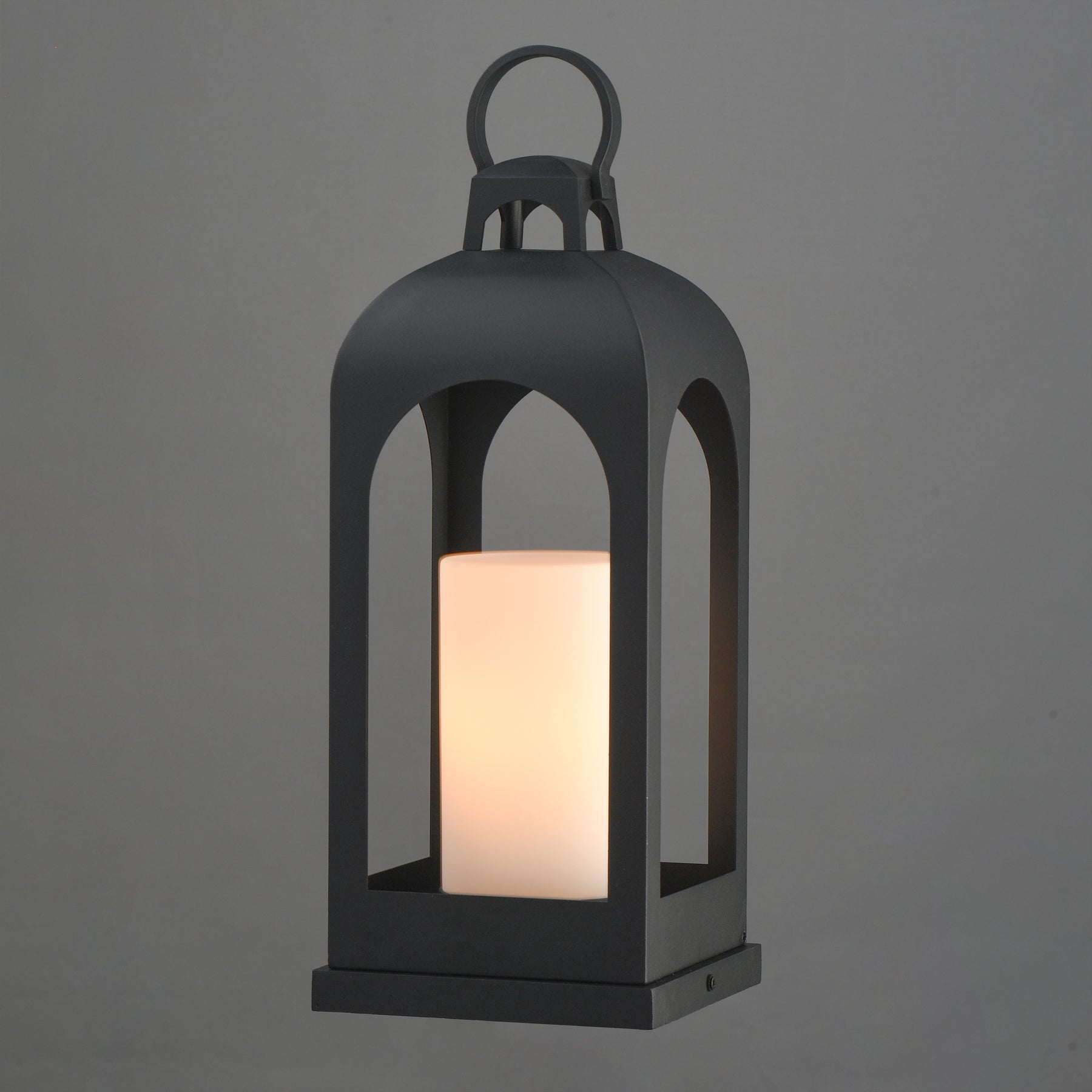 A black metal lantern-style outdoor light with a glowing warm white cylindrical candle inside, shown against a plain gray background.