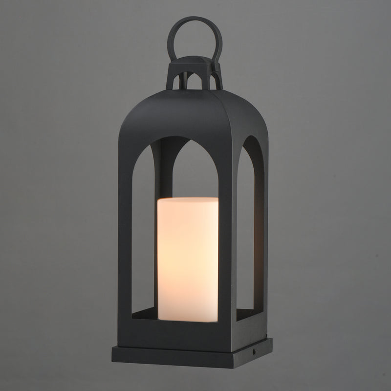 A black metal lantern-style outdoor light with a glowing warm white cylindrical candle inside, shown against a plain gray background.