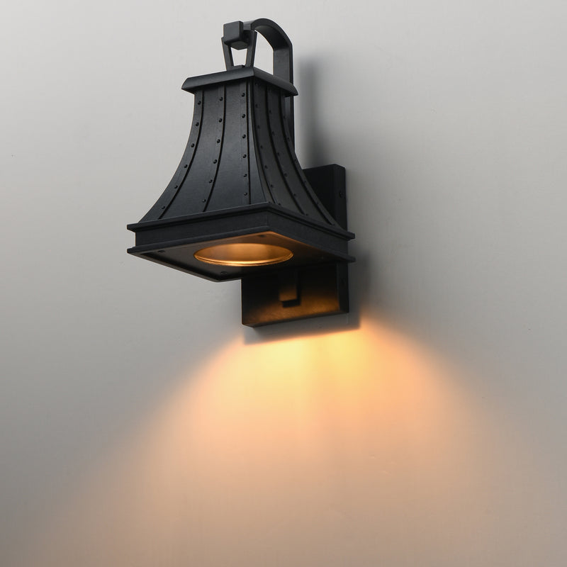 A black metal outdoor wall sconce with a warm yellow light glowing from within, mounted on a plain light gray wall.