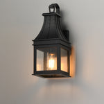 A black metal outdoor wall sconce with a warm glowing light inside, mounted on a plain light gray wall.