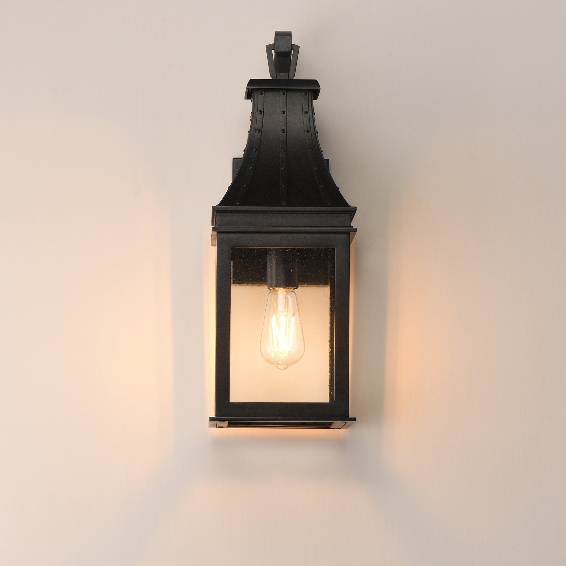 A black metal lantern-style wall sconce with a warm glowing Edison bulb, mounted on a plain off-white wall.