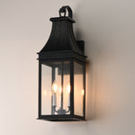 A black metal outdoor wall sconce with a lantern-style design, featuring clear glass panels and two visible lit candle-style bulbs, mounted on a plain light beige wall.