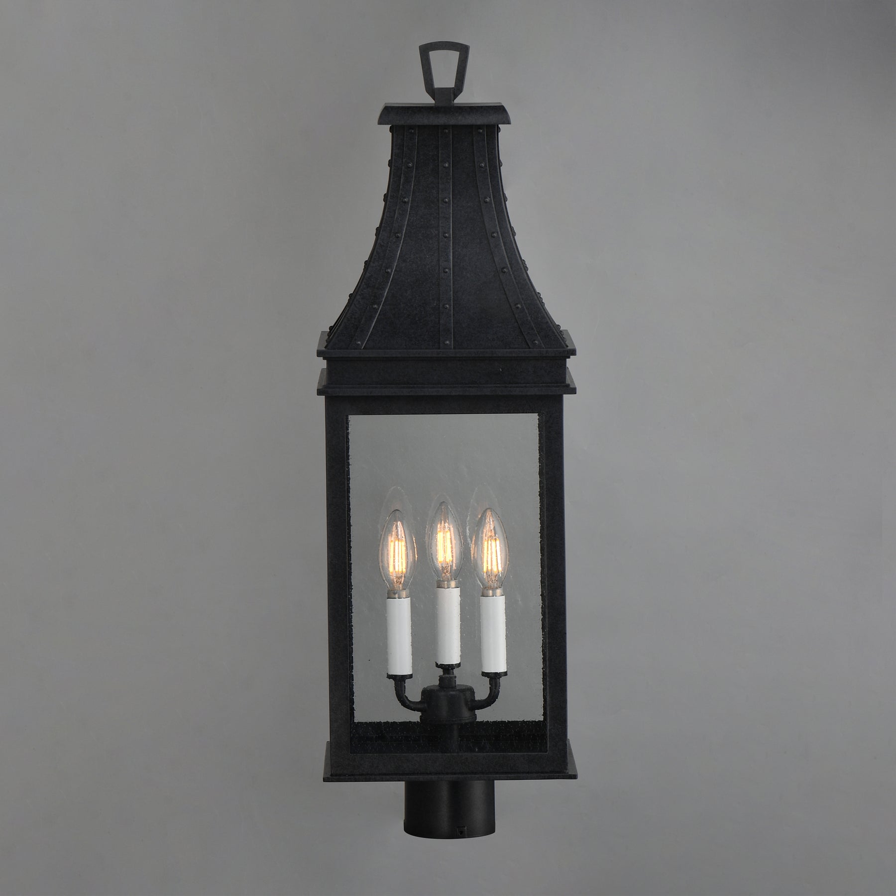 A black metal lantern-style wall light with three candle-style bulbs illuminated, shown against a plain gray background.