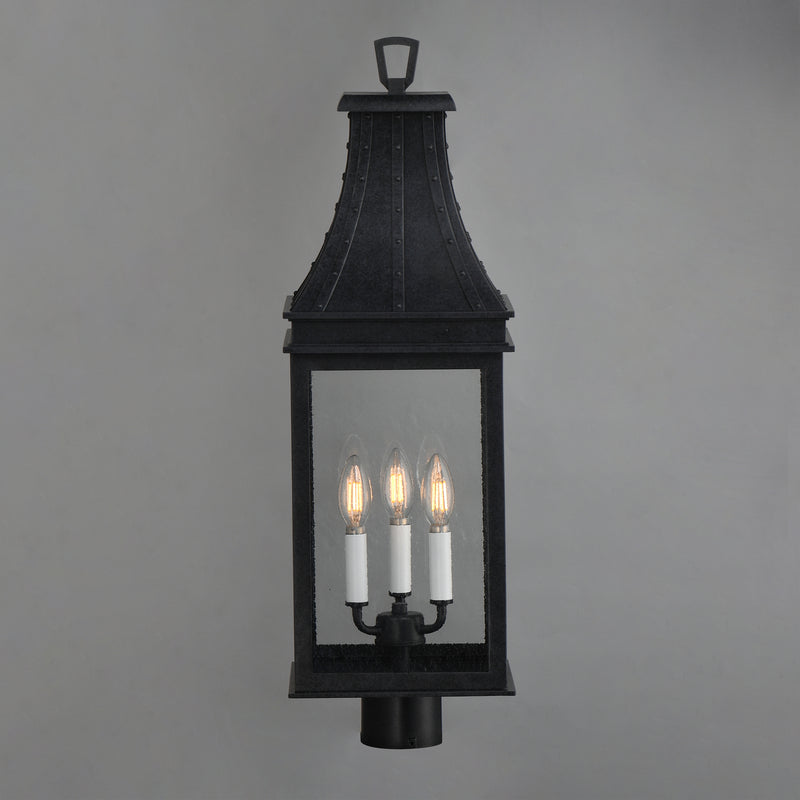 A black metal lantern-style wall light with three candle-style bulbs illuminated, shown against a plain gray background.