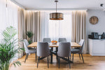 A modern dining room setup featuring a rectangular light wood dining table surrounded by six gray upholstered chairs with black legs. The room has beige curtains, a large brass drum pendant light, a wooden floor, and a small white side cabinet with a clock on the wall.