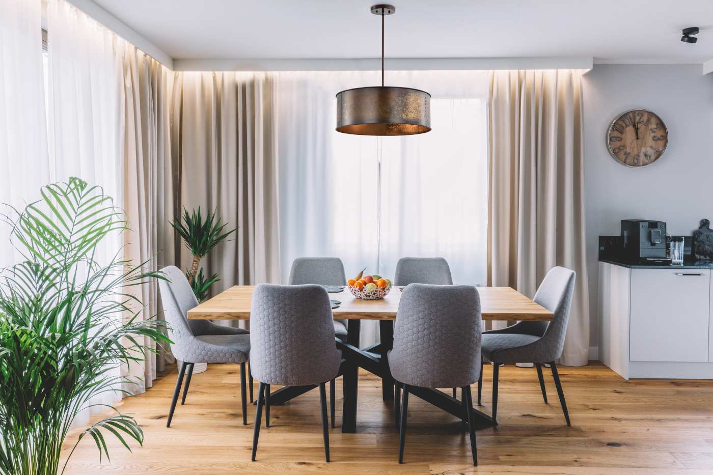 A modern dining room setup featuring a rectangular light wood dining table surrounded by six gray upholstered chairs with black legs. The room has beige curtains, a large brass drum pendant light, a wooden floor, and a small white side cabinet with a clock on the wall.