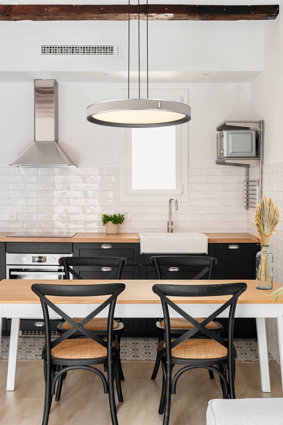 A modern kitchen and dining area featuring a light wood dining table with four black metal chairs that have woven cane seats, set against a white brick backsplash and black cabinets.