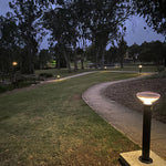 A modern black solar vandal resistant bollard light with a pink dome top emitting warm 588 lumens illumination