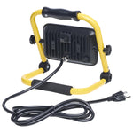 A portable work light with a yellow metal frame, black handle, and black cooling fins on the back, shown with its attached black power cord on a white background.