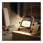 A yellow and black SATCO portable work light with a bright illuminated panel, shown on a stack of plywood in a construction site with tools, gloves, and scaffolding visible in the background.