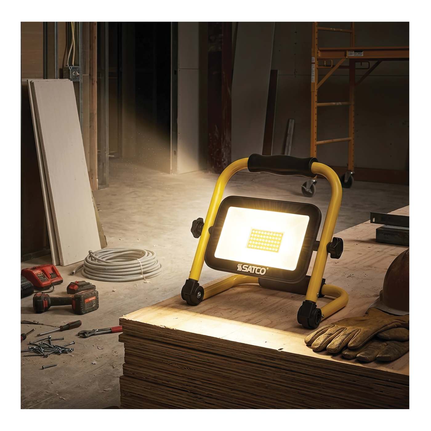 A yellow and black SATCO portable work light with a bright illuminated panel, shown on a stack of plywood in a construction site with tools, gloves, and scaffolding visible in the background.