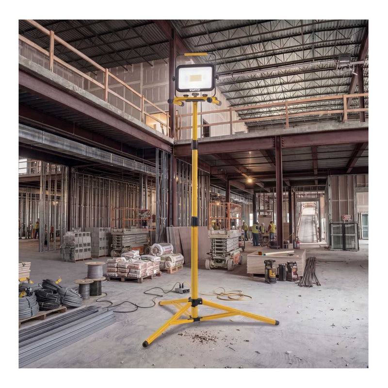 A yellow portable work light on a three-legged stand, positioned in the center of a large, active industrial warehouse or construction site.