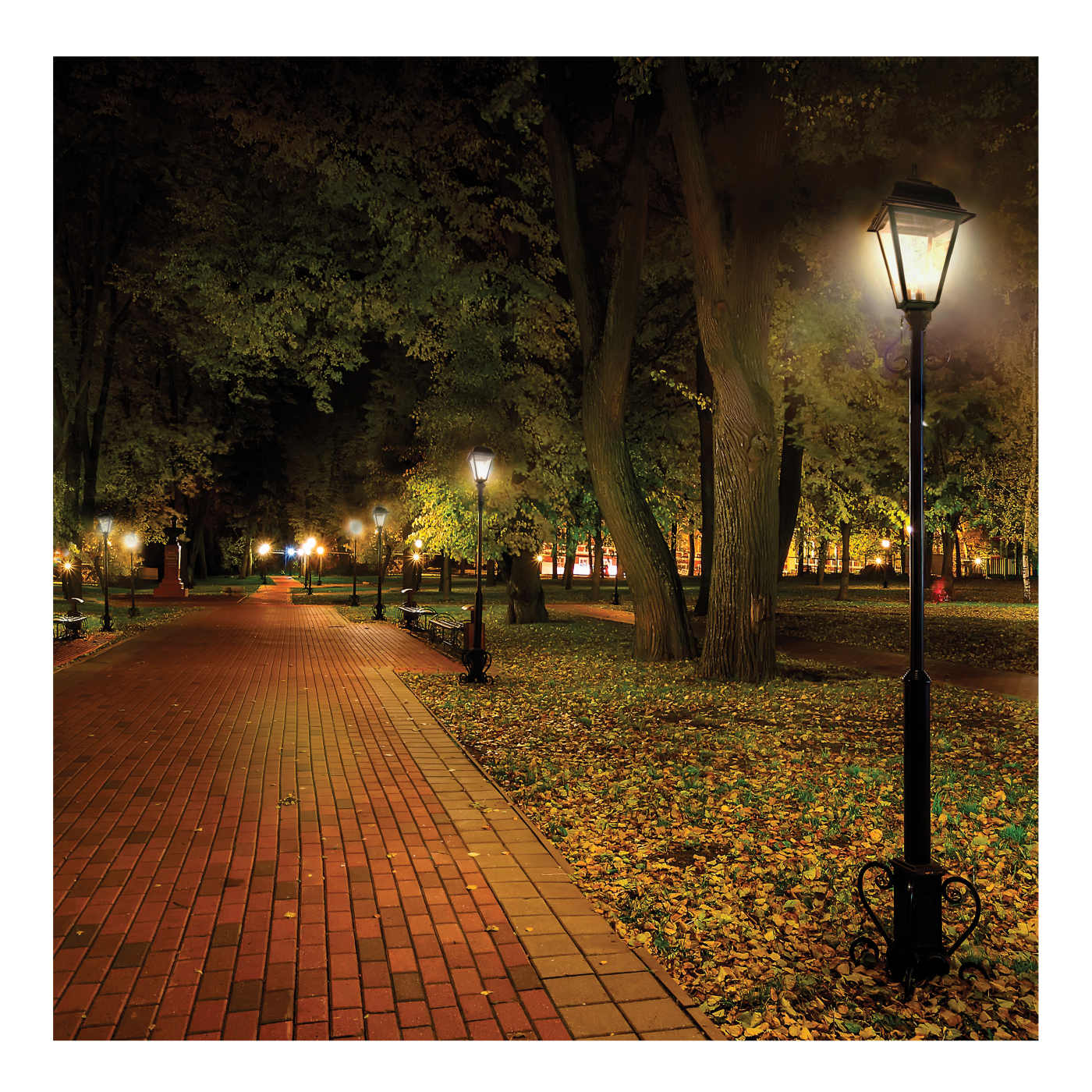 A classic black outdoor garden lamp post with a glowing warm light, situated on a leaf-covered lawn beside a brick pathway in a park at night.